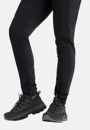 Jambes portant un pantalon noir slim et des chaussures de randonnée noires avec semelles grises et lacets à motifs sur un fond uni.