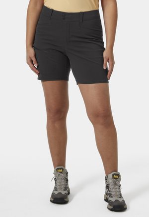 Vrouw die zwarte korte broek met ritszakken, een beige top en beige wandelschoenen draagt, staand tegen een witte achtergrond.