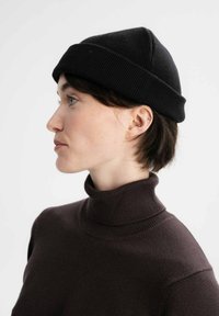 Zwarte gebreide beanie met een omgeslagen boord, gemaakt van zacht materiaal. Getoond op een model dat een donkerbruin coltrui draagt.