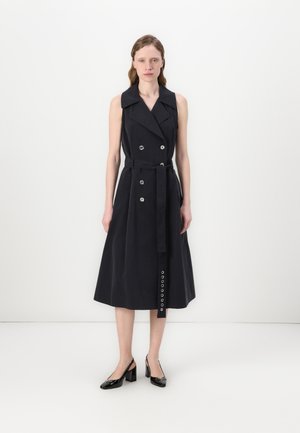 Laivastonsininen ilmanhihaista trench-mekko, jossa on kaksirivinen muotoilu, suuret napit, vyö ja levenevä helma; sileä kangas, joka muodostaa jämäkän siluetin.