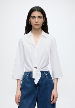 Frau mit langen dunklen Haaren, die goldene Creolen, ein weißes, geknöpftes Hemd, das an der Taille gebunden ist, und hoch taillierte blaue Jeans vor weißem Hintergrund trägt.