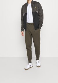 Chándal masculino que incluye una chaqueta con cremallera en gris oscuro y negro con bloques de color, combinado con joggers verdes oliva. Zapatillas blancas.