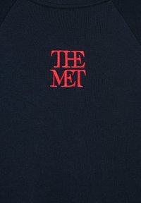 Testo ricamato rosso "THE MET" centrato su tessuto blu scuro, probabilmente una felpa o una t-shirt.