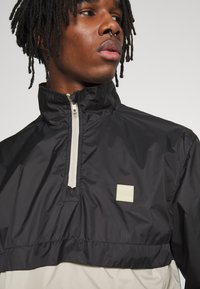 Giacca a vento nera e beige chiaro con colletto alto, zip frontale a metà e una toppa logo testurizzata sul petto. Tessuto liscio e leggero.
