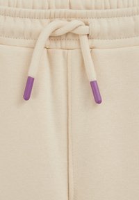 Bež trenirke z elastičnim pasom, z zvezanim vrvico s vijoličnimi konicami. Mehka tekstura in preprosti oblikovalski detajli.