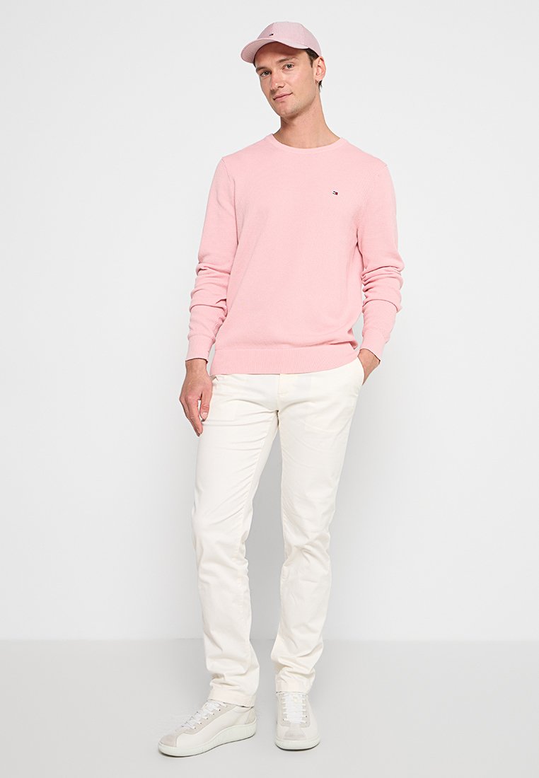 Helles rosa Strickpullover, weiße Hose und beige Turnschuhe. Der Pullover hat ein kleines Logo auf der Brust und einen runden Ausschnitt.