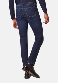 Jean slim bleu foncé avec cinq poches, couture visible et étiquette de marque à la taille. Accompagné de bottines noires.