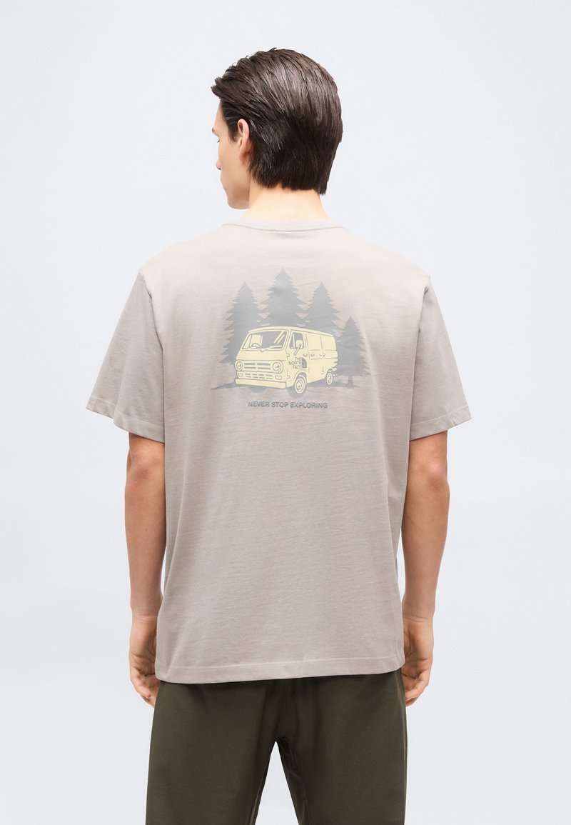 Man wearing beige T-shirt with a yellow van and pine trees graphic and "Never Stop Exploring" text on the back, standing against a plain background.