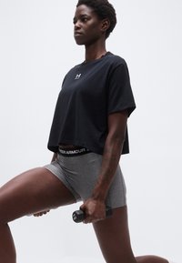 Γυναίκα με μαύρο κοντομάνικο μπλουζάκι και γκρι σορτς κρατάει ένα αλτήρα 1 κιλού ενώ εκτελεί άσκηση προβολής σε απλό φόντο.