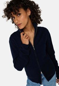 Pull-over zippé marine avec un tricot texturé, col haut et ourlet festonné. Il arbore des manches longues et une quincaillerie en métal argenté pour la fermeture éclair.