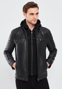 Chaqueta de motorista de cuero negra con hombros acolchados, dos bolsillos de pecho con cremallera y un diseño ajustado sobre una sudadera negra con capucha. Textura suave.