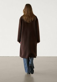 Brauner Wollmantel mit lockerer Passform, langen Ärmeln und gerader Silhouette. Über blauen Schlagjeans getragen, von hinten betrachtet.