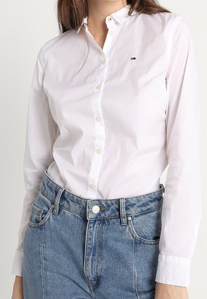 Camisa blanca de botones con mangas largas, cuello clásico y pequeño logo. Combinada con jeans de mezclilla azul de cintura alta.