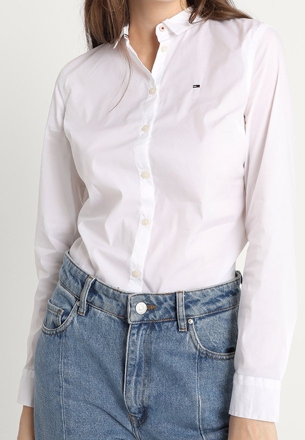 Camisa blanca de botones con mangas largas, cuello clásico y pequeño logo. Combinada con jeans de mezclilla azul de cintura alta.