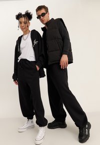 To unge voksne iført sort afslappet tøj og sneakers poserer mod en ensfarvet baggrund, den ene med solbriller og en dunvest.