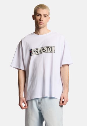 Młody mężczyzna z krótkimi blond włosami, ubrany w oversize'ową białą koszulkę z napisem „PROSTO” oraz jasnoniebieskie dżinsy, stojący na jednolitym tle.