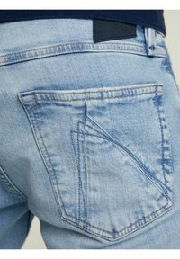 Los vaqueros de mezclilla azul claro presentan un bolsillo trasero distintivo con un diseño angular, costuras visibles y un parche de cuero negro cerca de la cintura.