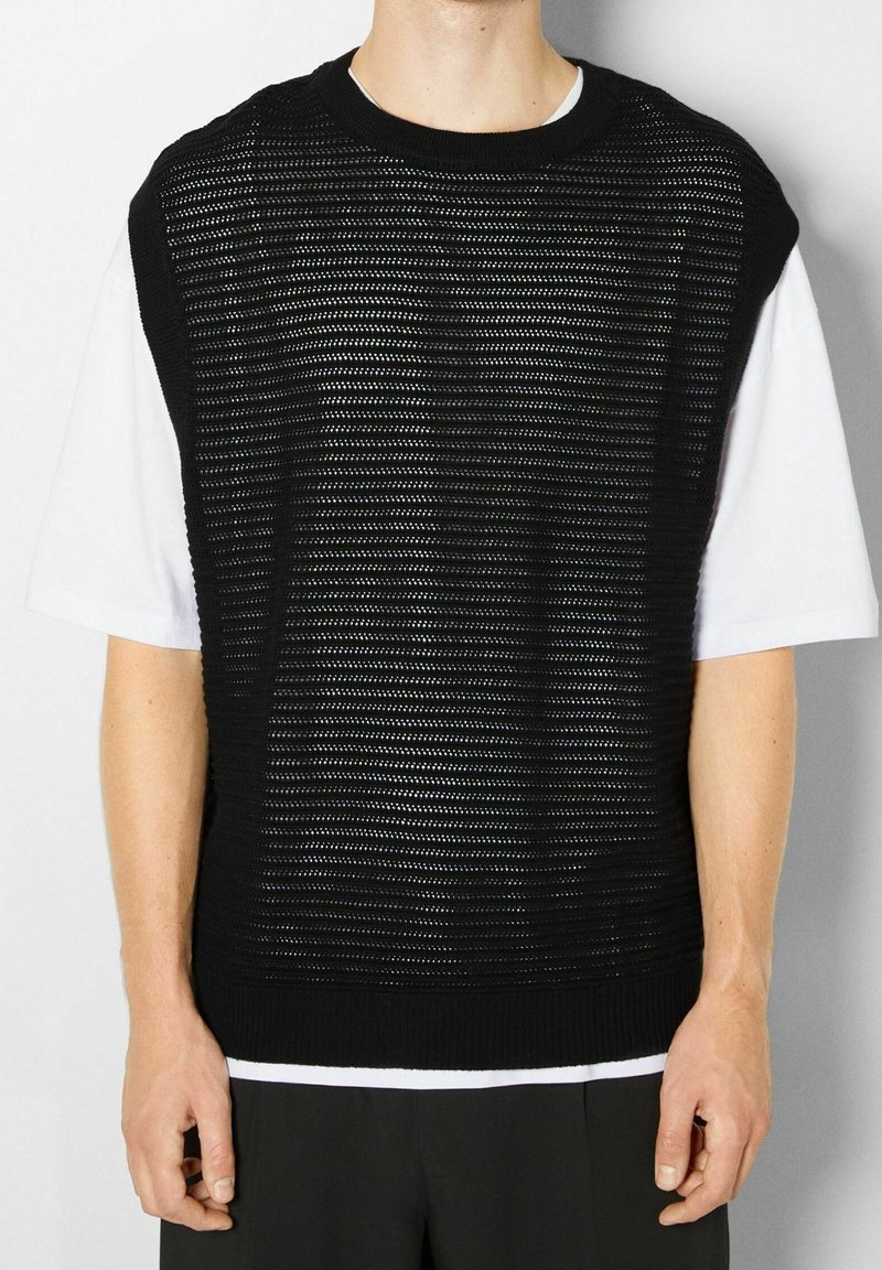 Haut sans manches en maille noire à rayures horizontales, superposé sur une chemise blanche à manches courtes. Tissu texturé et col rond.