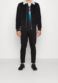 Svart kordjacka med en vit fuskpälskrage, bärs över en ombre grafisk t-shirt, ihop med svarta byxor och svarta sneakers.