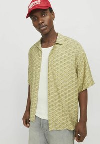 Grünes Kurzarmshirt mit geometrischem Muster, aus leichtem Stoff. Getragen über einem weißen gerippten Tanktop.