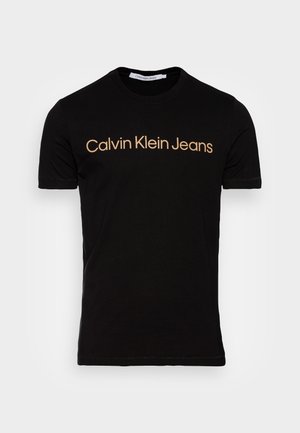 Czarny T-shirt z krótkim rękawem i okrągłym dekoltem, z beżowym napisem "Calvin Klein Jeans" na środku klatki piersiowej.
