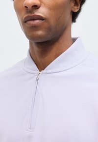 Κοντινό πλάνο του κάτω μέρους του προσώπου και του λαιμού ενός ατόμου που φοράει ένα ανοιχτό μοβ πουκάμισο με φερμουάρ, με το φερμουάρ μερικώς κλειστό.