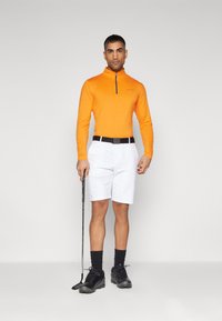 Oranžna dolg rokav polo majica z zadrgo, bele kratke hlače s žepi, črn pas, črne nogavice in črne golf čevlje, drži golf palico.