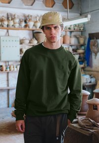 Sudadera verde oliva con cuello redondo y puños acanalados, con un sutil detalle de logotipo. El modelo lleva una gorra beige y está en un taller de cerámica.