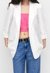 Weißer Blazer mit aufgerollten Ärmeln, der ein maßgeschneidertes Design hat. Kombiniert mit einem rosa Cropped-Top und hochgeschnittenen blauen Jeans mit einem schwarzen Gürtel.