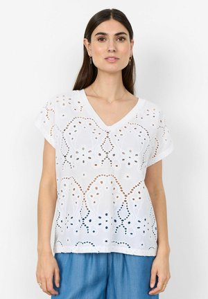 Frau trägt eine weiße Bluse mit Kurzarm und Lochstickerei-Muster sowie V-Ausschnitt und blaue Hose, steht vor einem schlichten Hintergrund.