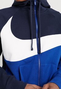 Námořnická a modrá mikina na zip s bílým designem ve tvaru šmouhy. Má klokánkovou kapsu a kapuci se stahovací šňůrkou. Měkká textura látky.