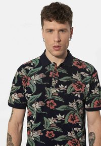 Zwarte polo met bloemmotief, versierd met rode en groene bloemen. Het heeft een klassieke kraag en een drieknoopsluiting, en is gemaakt van gestructureerde stof.