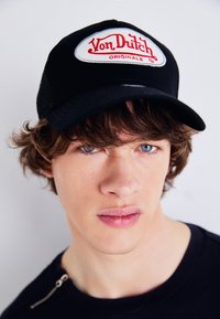 Zwarte trucker pet met een witte geborduurde patch met de tekst "Von Dutch" en "ORIGINALS" in het rood. Model draagt een donker shirt met een ritsdetail.