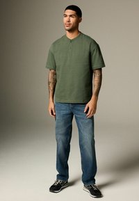 Olive green short-sleeve henley shirt with three buttons, paired with light blue relaxed-fit jeans and black running shoes.