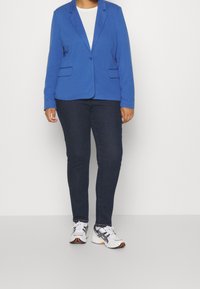 Niebieska marynarka z wyciętym kołnierzem, dwoma przednimi kieszeniami, w zestawieniu z ciemnymi jeansami i białymi trampkami. Gładka tkanina, dopasowany fason i casualowy styl.
