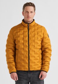 Gelbe Pufferjacke mit gestepptem Textil, hohem Kragen und Reißverschluss. Besitzt seitliche Taschen und schwarze Akzente für einen Kontrast.