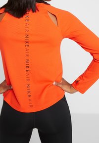 Orange langærmet sportstrøje med struktureret mønster, udsparinger ved skuldrene og sort "NIKE AIR" branding midt på ryggen.