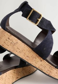 Sandale din suede bleu marin, cu platformă din plută, având o curea lată la gleznă și un detaliu cu cataramă aurie. Talpa este maro și ușor conturată.