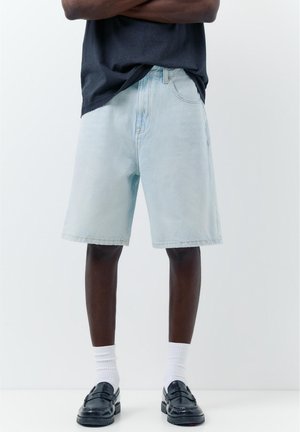 Férfi világoskék farmer rövidnadrágban, fehér bokazokniban, fekete loaferekben és sötét ingben, karját keresztbe téve, egyszínű háttér előtt áll.