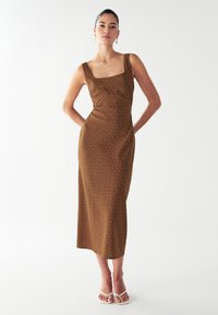 Vestido midi marrón con escote cuadrado, sutiles lunares blancos, corsé ajustado y falda recta. La modelo posa con las manos en los bolsillos.