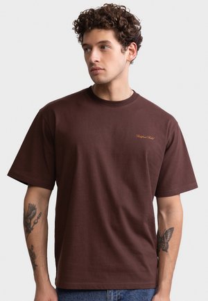 Ung mand med krøllet hår iført en mørkebrun T-shirt med lille orange tekst på brystet, der viser tatoveringer på begge underarme.