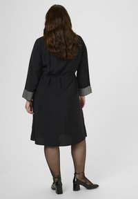 Vrouw met lang bruin haar staat met haar rug naar je toe, gekleed in een zwarte jurk tot aan de knie met zilverkleurige studs op de manchetten en zwarte gepatineerde hakken met enkelbandjes.