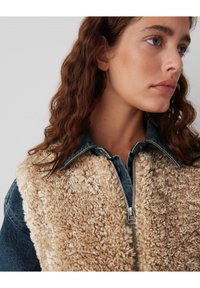 Veste en denim avec un doublure en shearling beige et duveteux. Dispose d'une fermeture éclair à l'avant et d'un col, mettant en valeur des textures et des teintes contrastées.