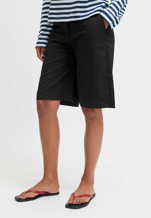 Person trägt schwarze knielange Shorts, ein blau-weiß gestreiftes Langarmshirt und rote Flip-Flops und steht auf weißem Hintergrund.