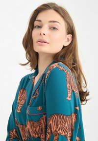 Teal blouse met een overal tijgerprint in oranje, voorzien van een v-hals, een zak en contrasterende rode piping langs de kraag.