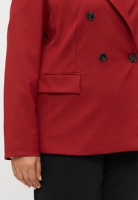 Raudona dvigubo užsegimo švarkas, pagamintas iš lygaus audinio. Yra du juodi mygtukai, krūtines kišenė ir šoninė nuovargio kišenė. Priklausomai nuo dizaino.