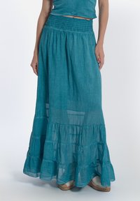 Una lunga gonna a strati realizzata in tessuto leggero color teal, con una vita arricciata e strati trasparenti, abbinata a scarpe espadrillas.