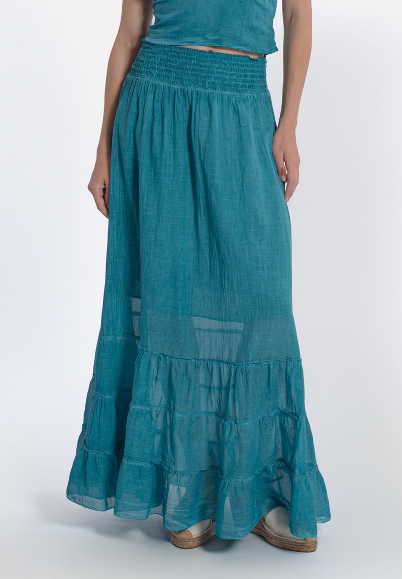 Una lunga gonna a strati realizzata in tessuto leggero color teal, con una vita arricciata e strati trasparenti, abbinata a scarpe espadrillas.