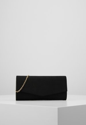 Pochette rectangulaire noire avec une lanière chaîne dorée posée sur une surface blanche contre un fond gris uni.