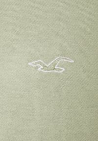 Tejido de punto verde claro con un acabado texturizado; presenta un logo de gaviota bordado en blanco situado a la izquierda.